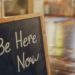 'Be Here Now' sign on board as in a yoga room.