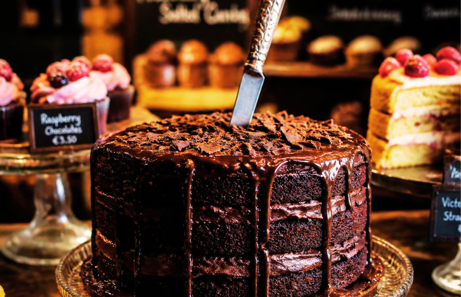 A dark mysterious layered fudge cake with a knife in it, dead center, no candles in a cake shop with other cakes around.