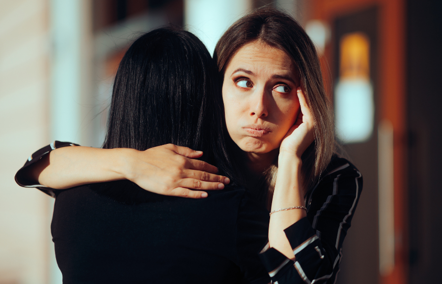 Image of two women hugging but one is facing forward and scrunching her face in judgement, not genuine.