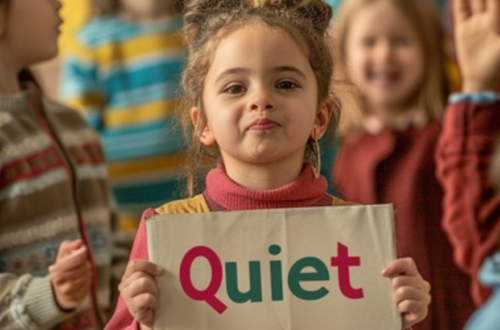 Young girl about five years old in the middle of a blurred image of her peers, she is resolute, holding a sign that reads 'Quiet'.
